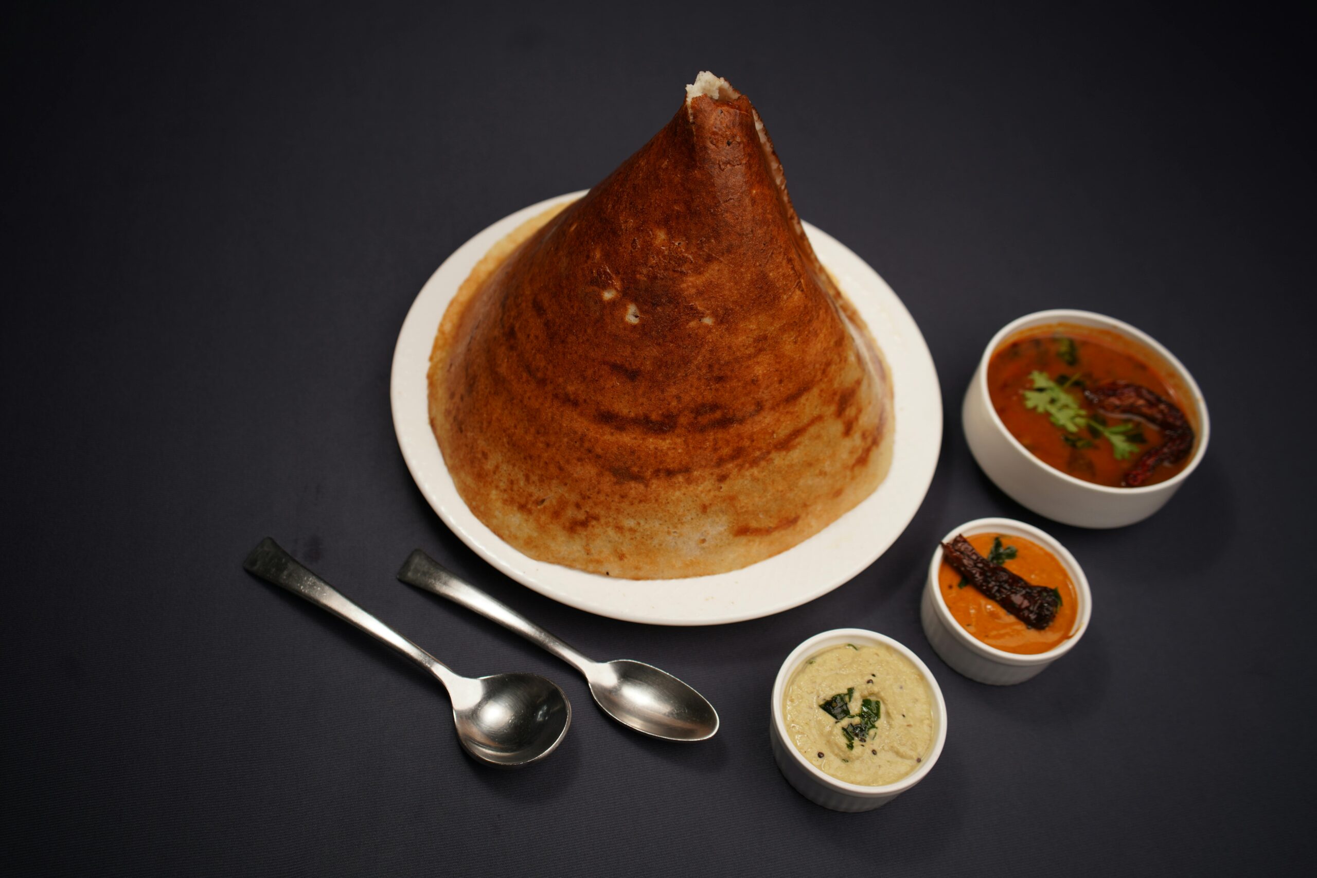 Crispy cone dosa served with authentic Indian chutneys and sambar on a dark backdrop.