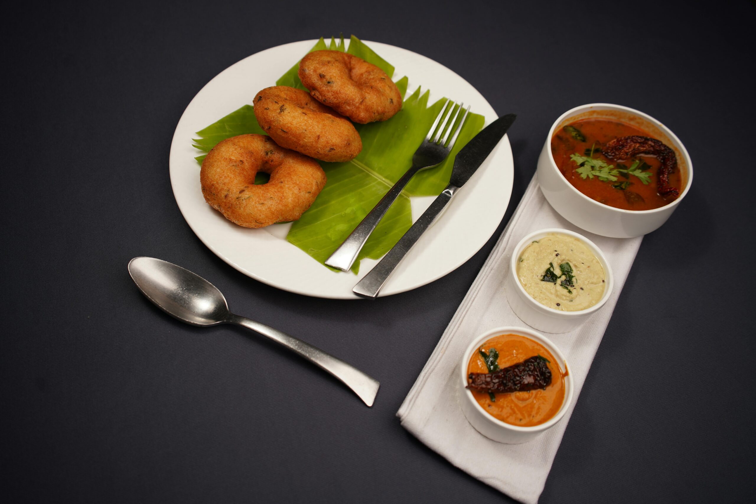A classic South Indian breakfast featuring vadai served with sambar and chutneys on a black background.